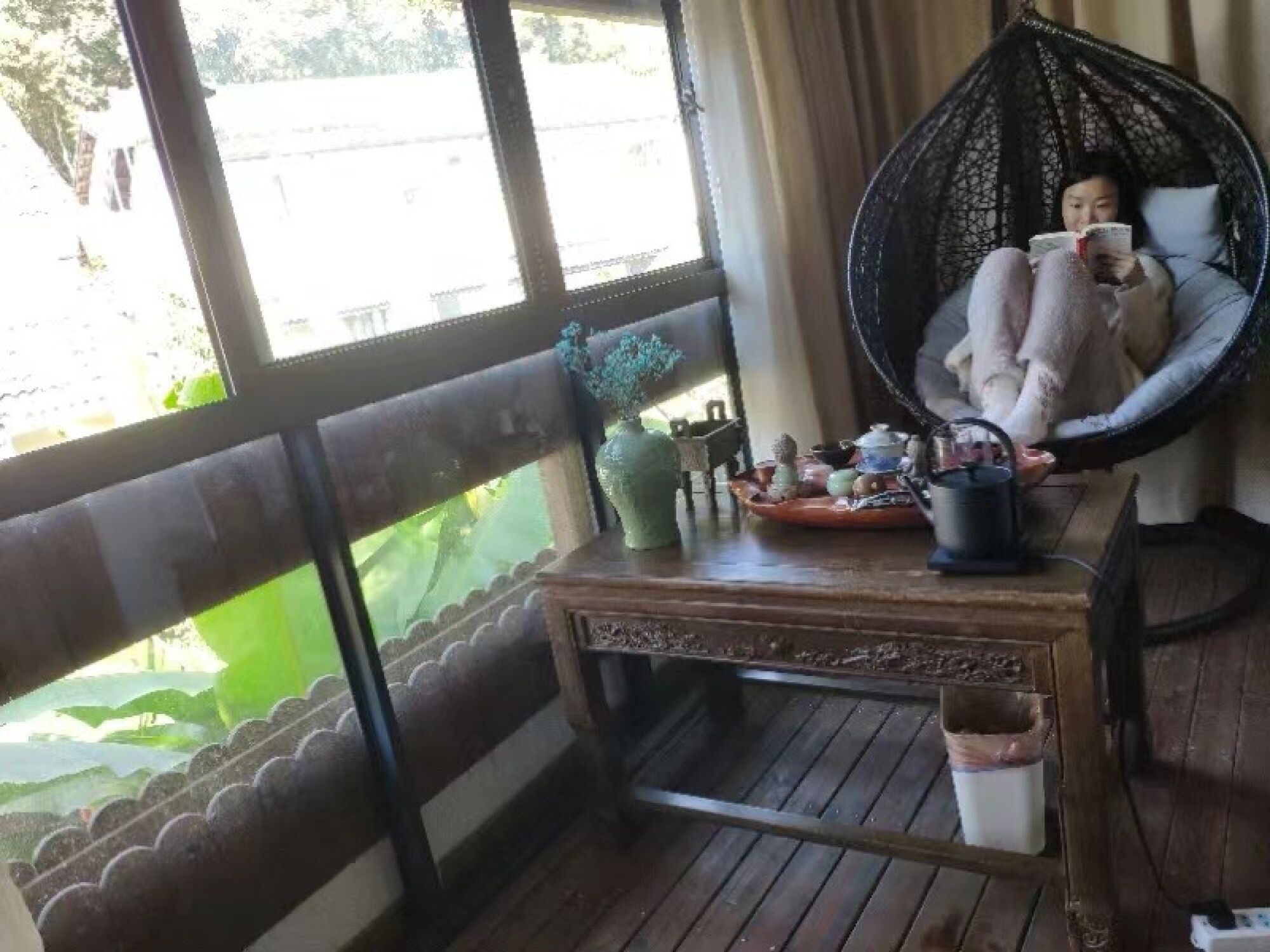 A woman sits reading a book in a large chair along a row of windows 