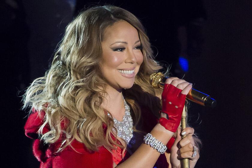Mariah Carey smiles while gripping a golden mic stand and wearing a red formal dress and matching gloves.