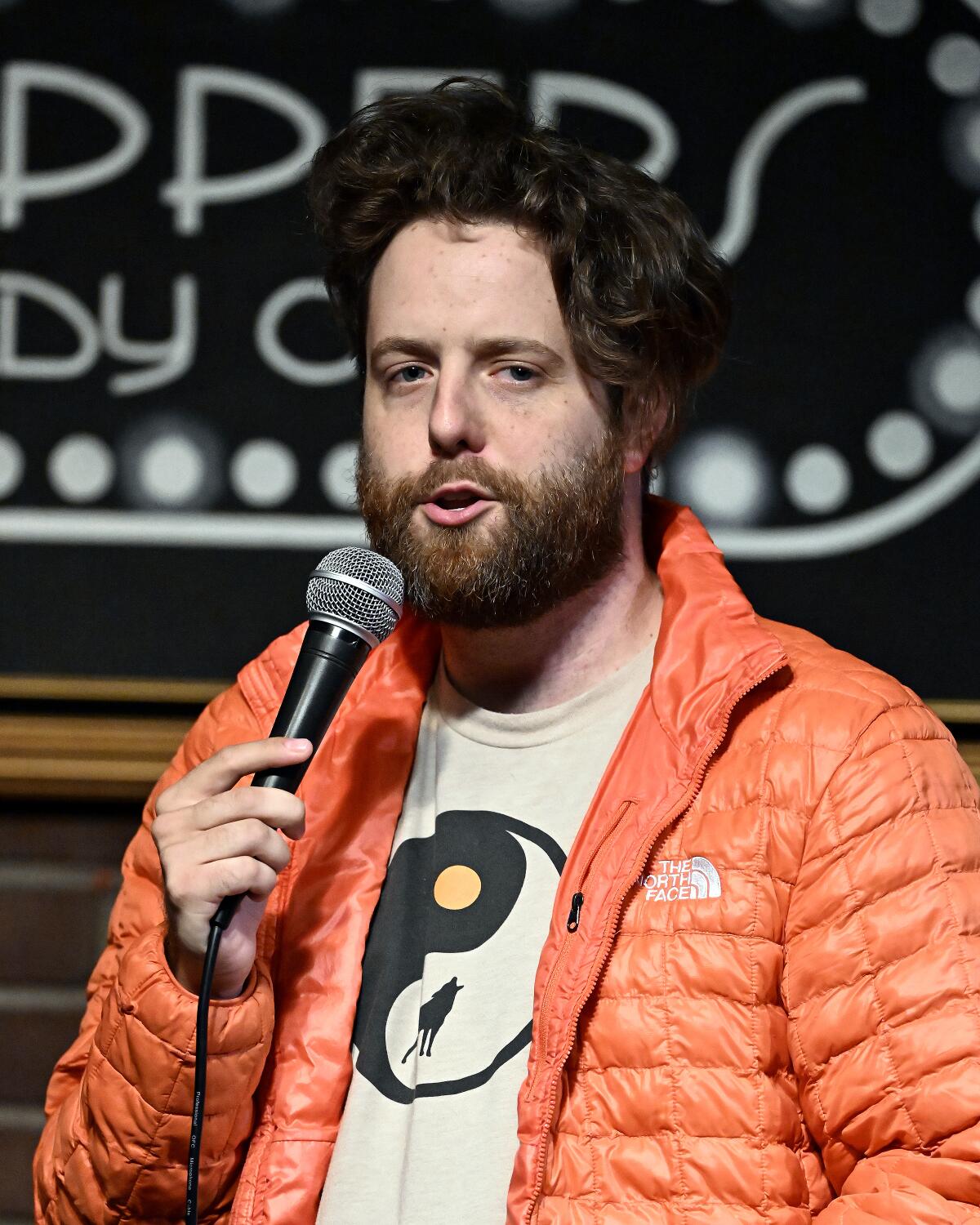 A bearded man in an orange puffer jacket speaks into a microphone