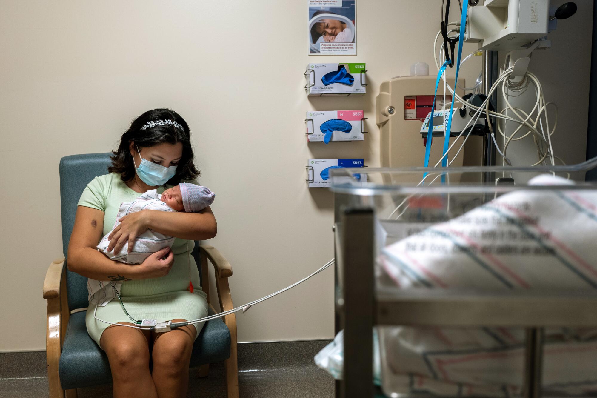 A woman with short dark hair, seated in a chair, holds a swaddled baby with a hat in a hospital room