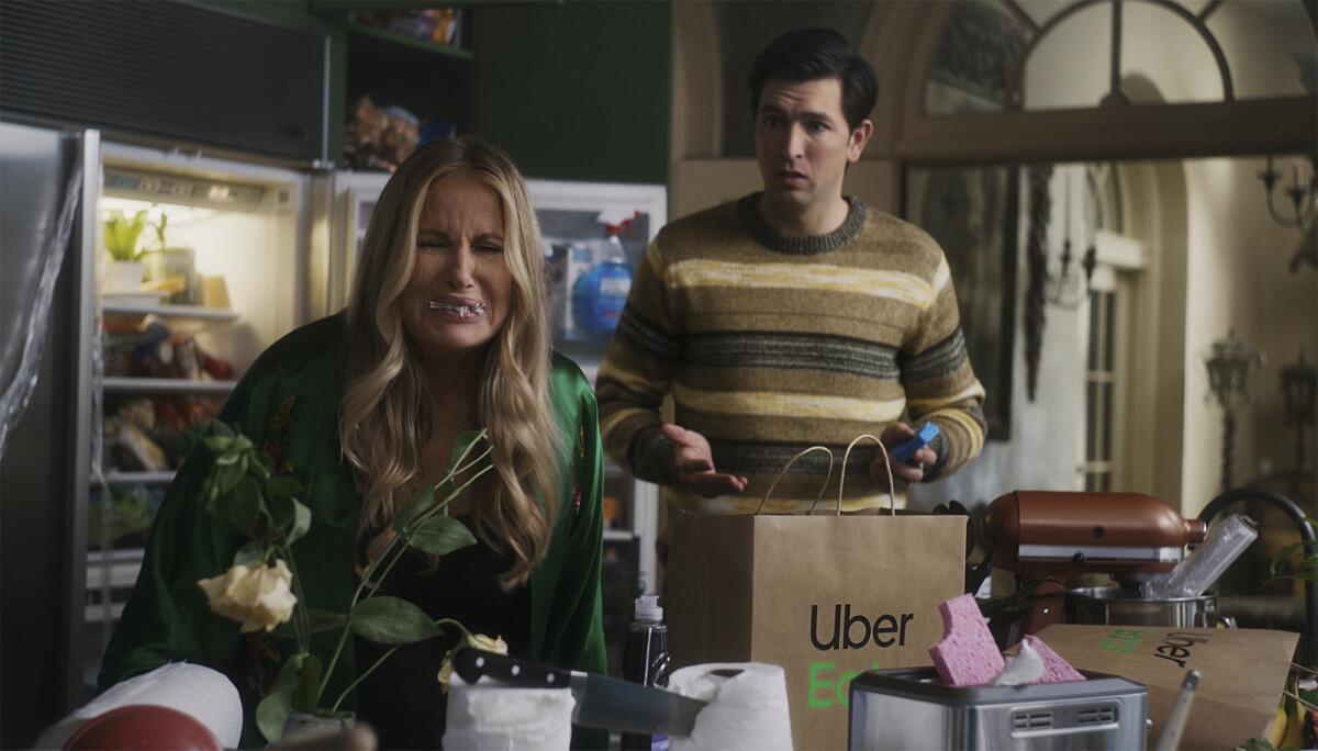 A woman with a mouth full of food stands next to a man with an Uber Eats bag in front of an open refrigerator.