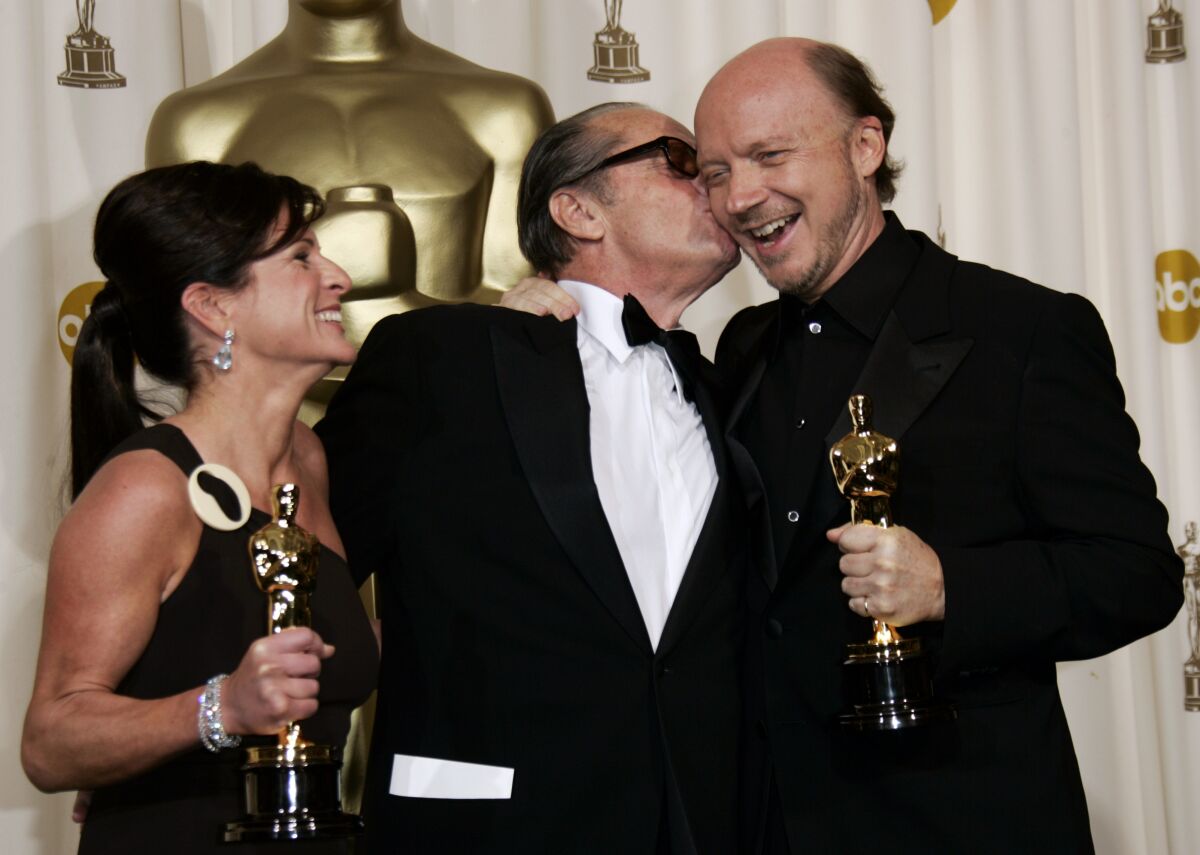 A man and a woman each hold award statues while a guy between them kisses the man on the cheek