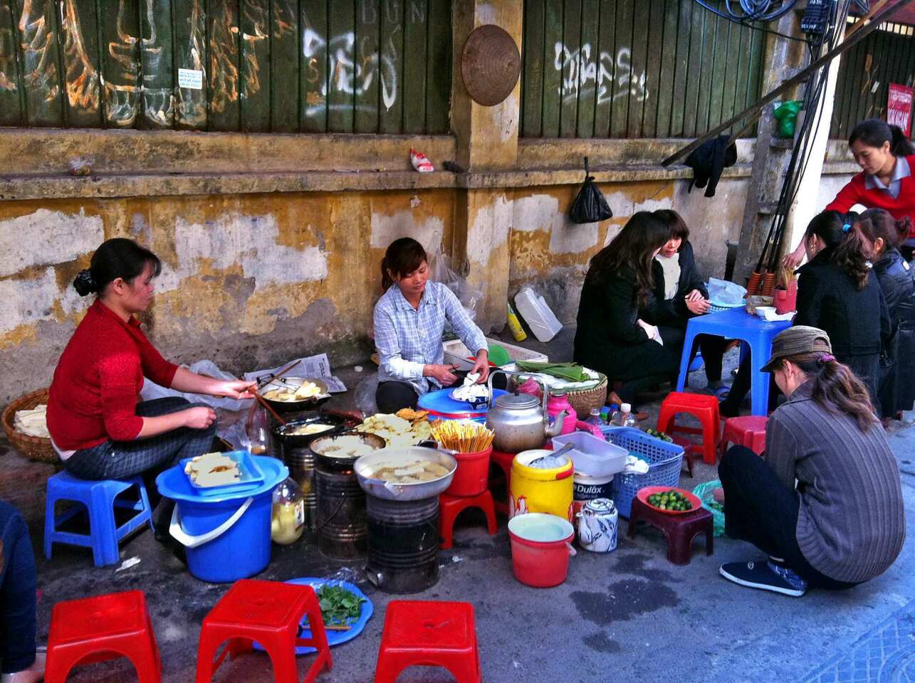 Women prepare a variety of soups, with a focus on bun dau (fried tofu soup), in an alley near the Hanoi Opera House. Broth is kept warm over coals in tin cans and diners squat on small plastic stools to consume their food.