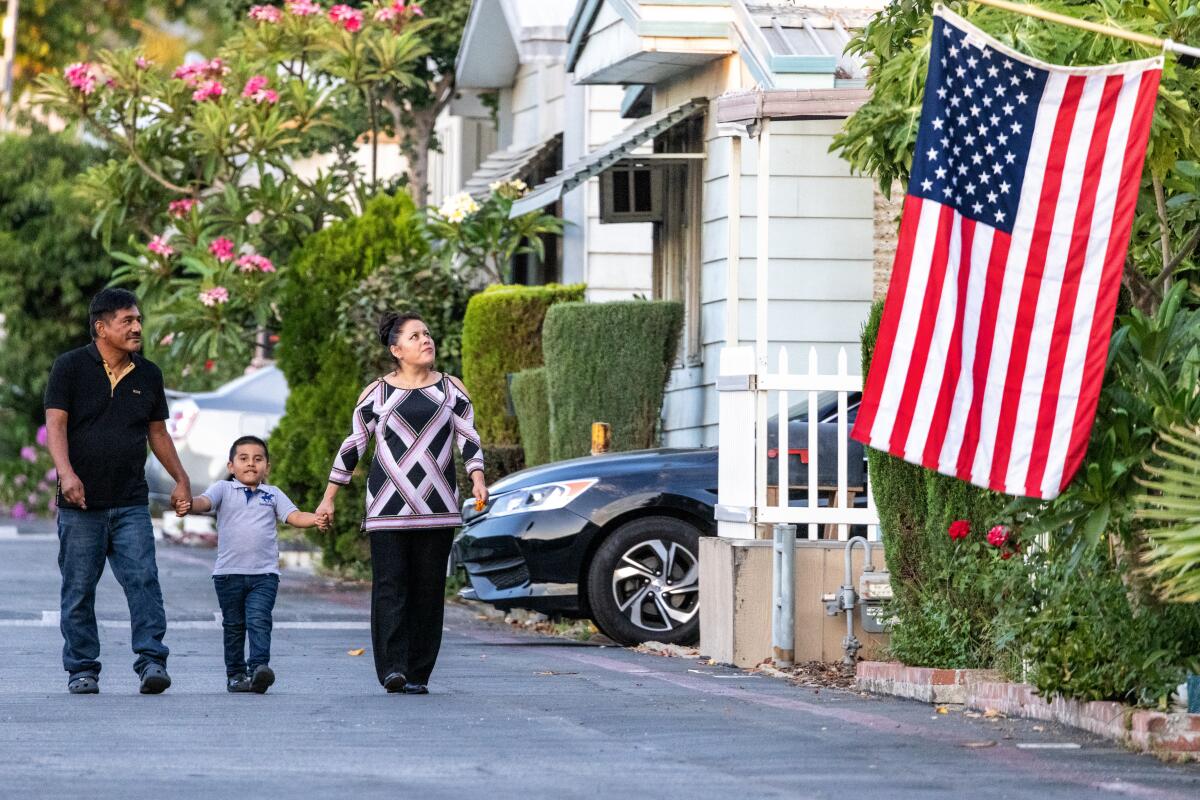 A boy holds hands with a man and a woman as they walk past homes, one of which has a U.S. flag