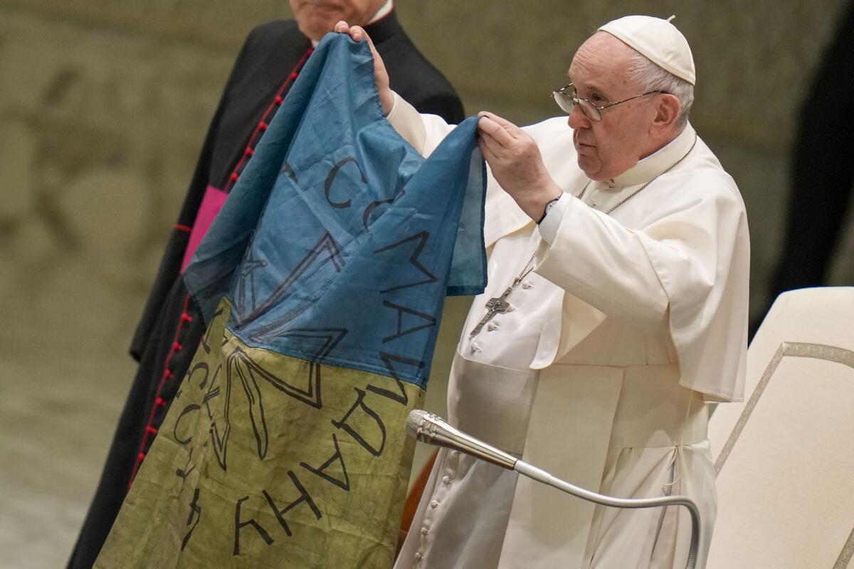 A man wearing glasses, a white cap and vestments holds up a blue and yellow flag 