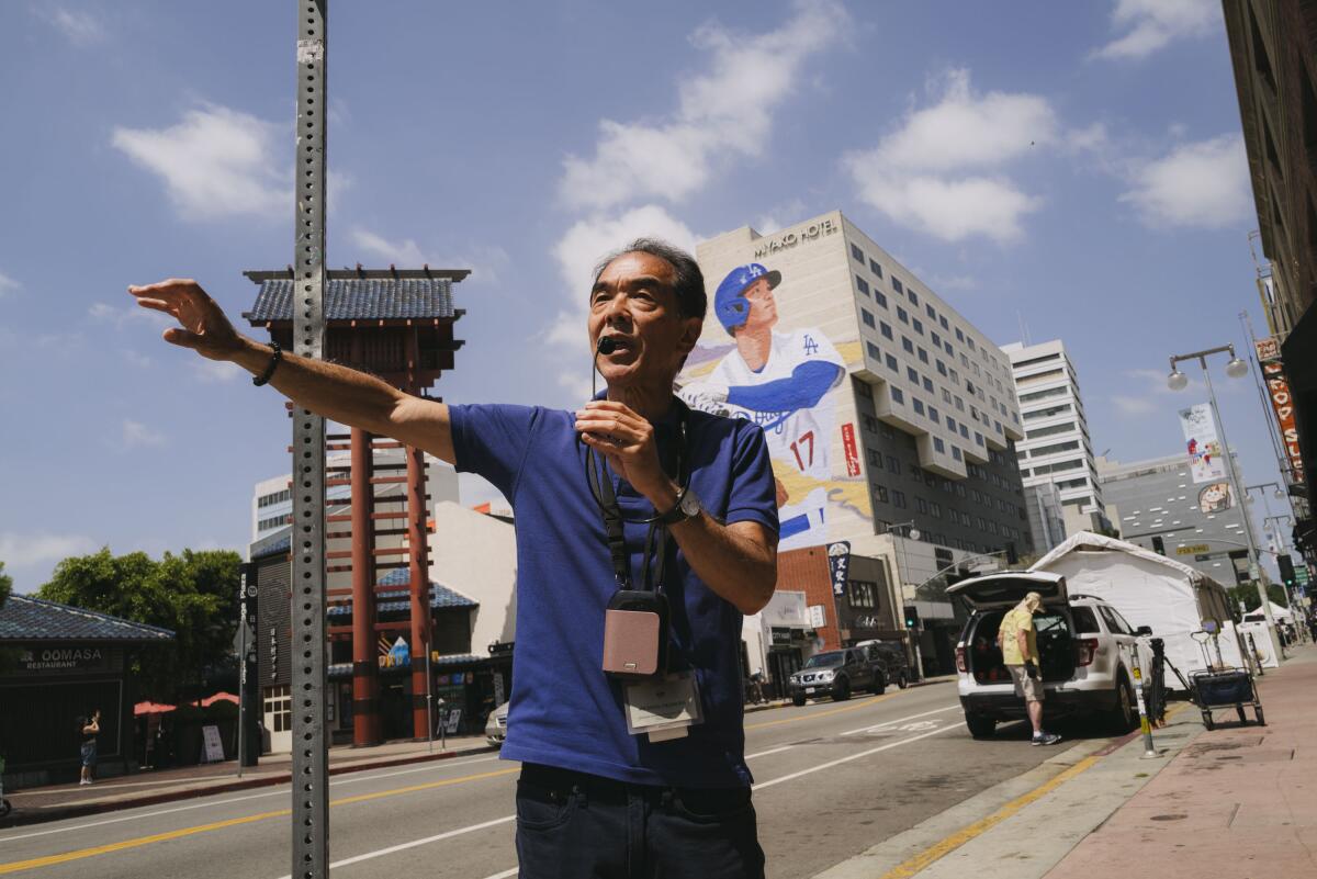 Michael Okamura holds a microphone and stands on a sidewalk with buildings in the background.