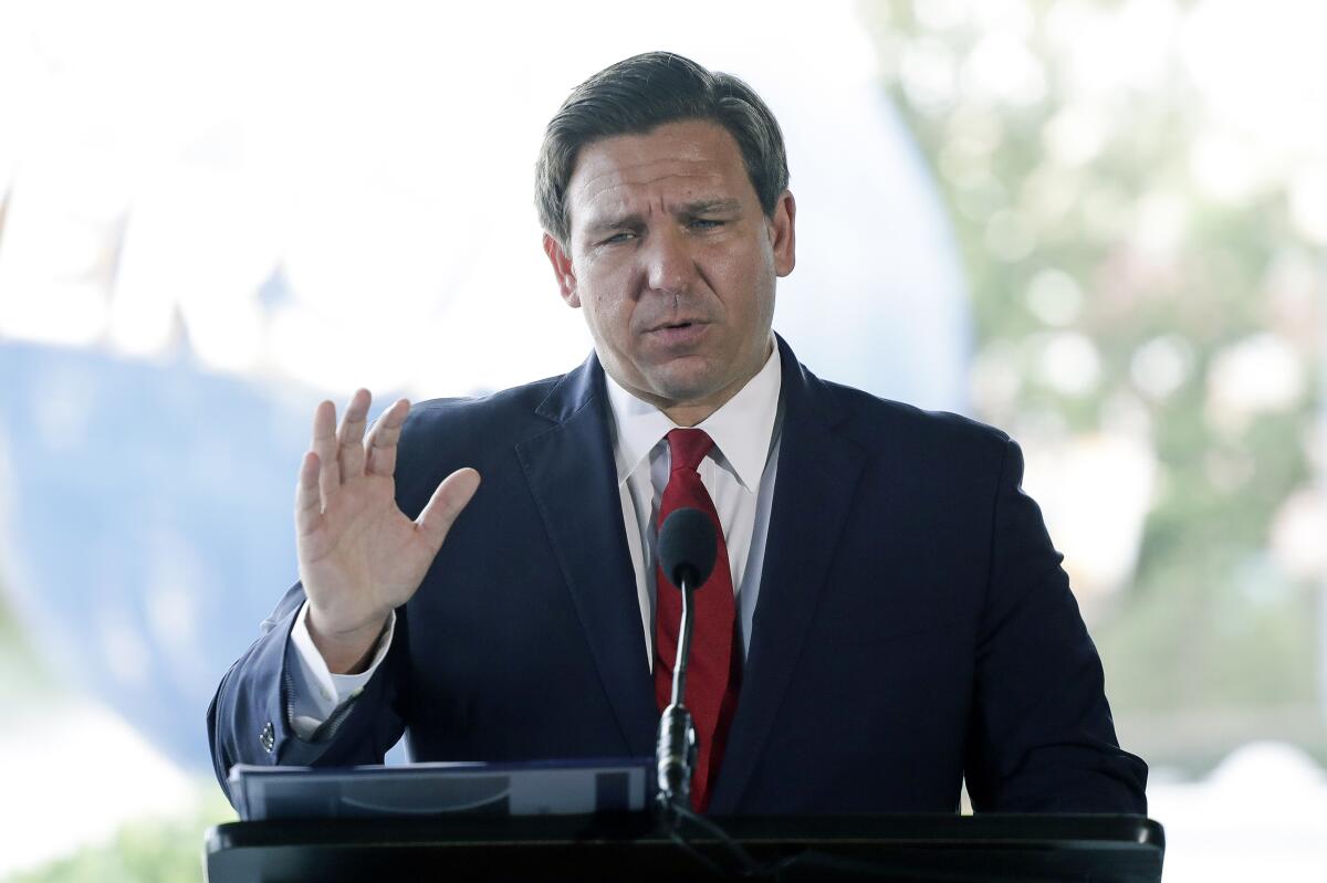 A man in a navy suit and red tie raises a hand as he speaks into a microphone at a podium.