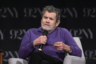A man with salt and pepper hair sitting in a gray lounge chair in a purple sweater holding a microphone and speaking