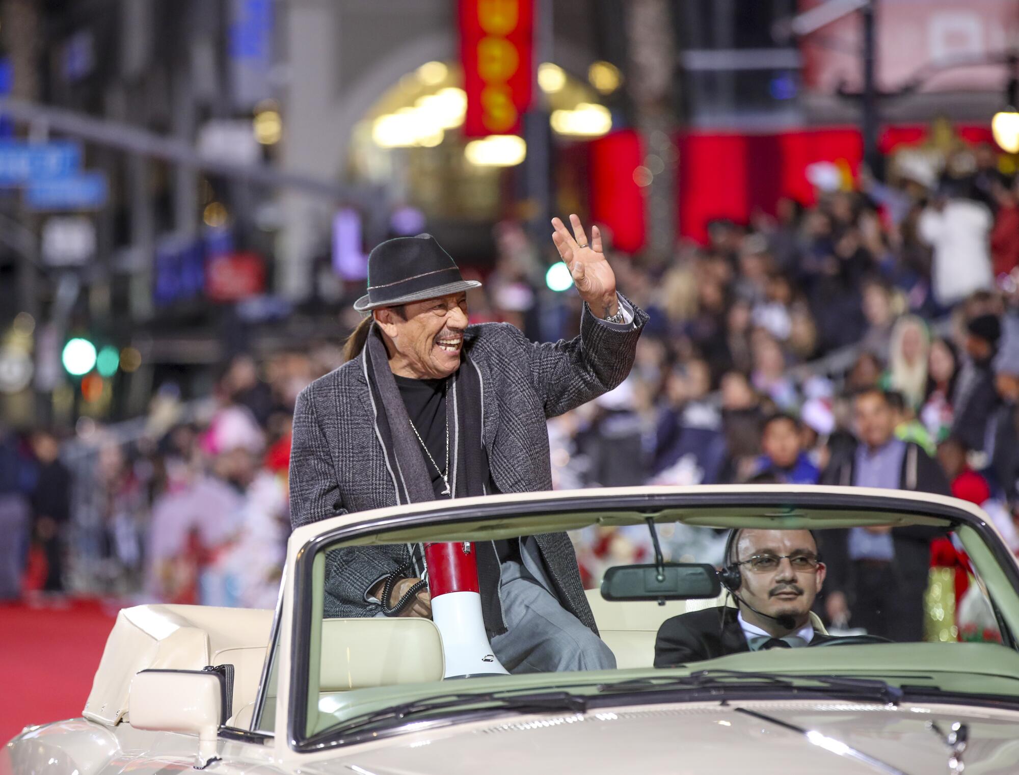 Actor Danny Trejo waves while sitting high on the backseat of a car while people watch in the background.