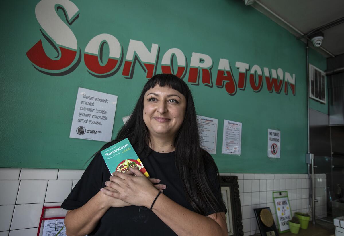  A smiling woman holds a book. Behind her is a green wall with the word "Sonoratown" in red and white.