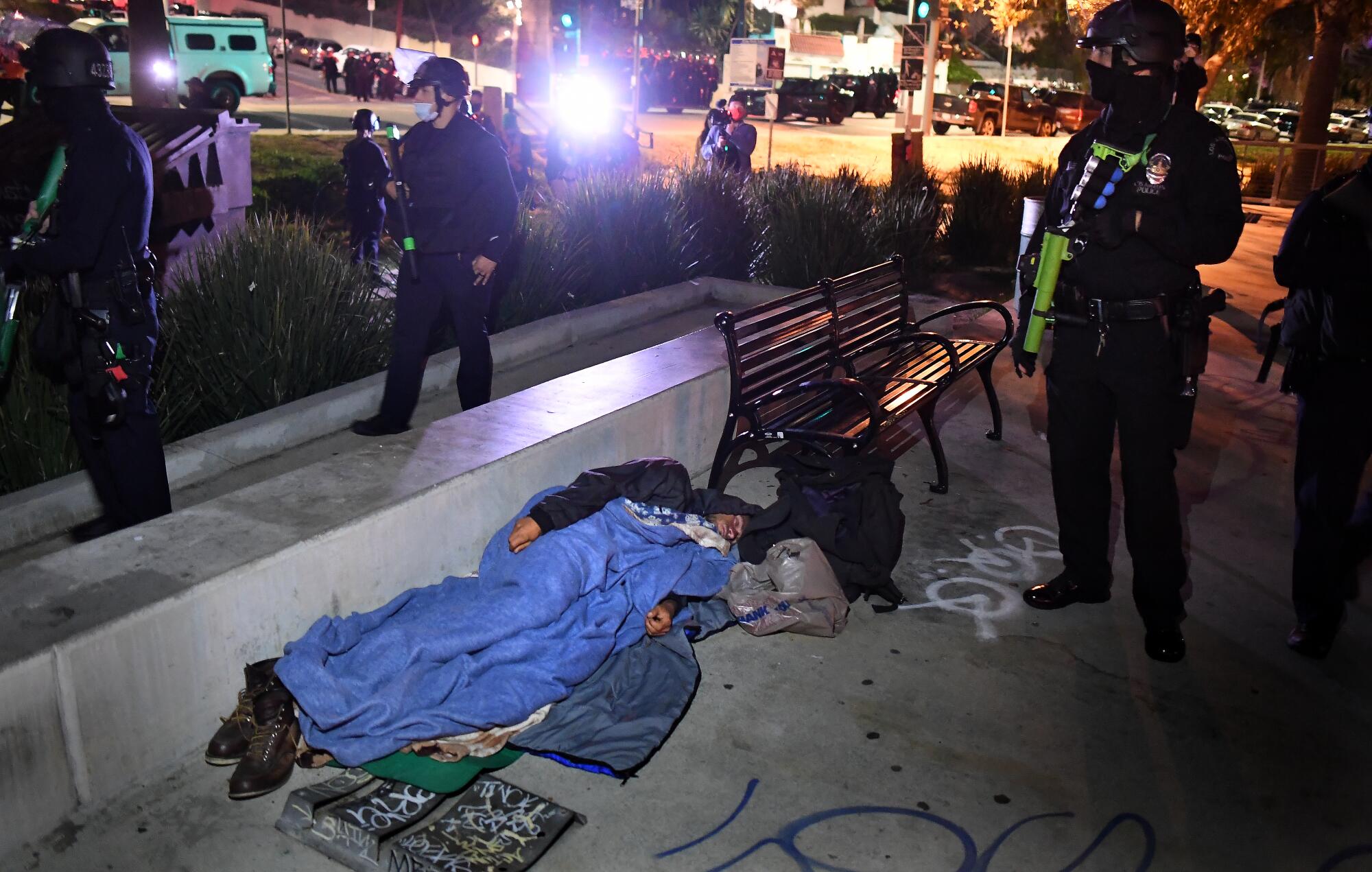 A person sleeps on the ground as police walk by.