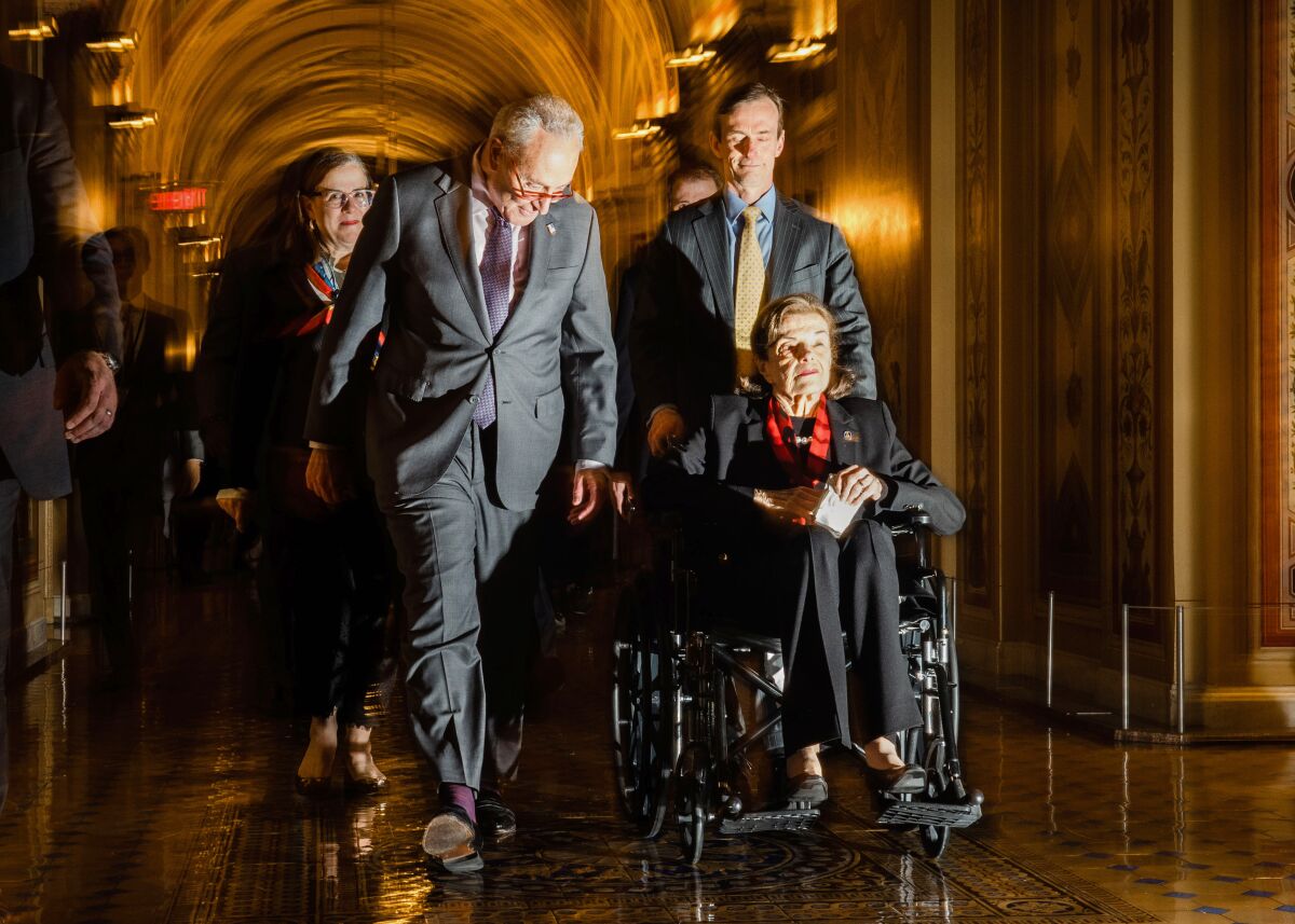 A person pushes another person in a wheelchair in an ornate hallway.