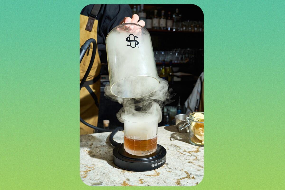 A glass container is lifted from over a cocktail glass. Smoke swirls around.