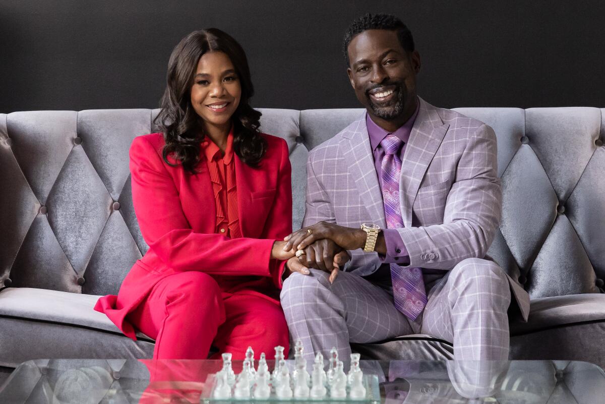 A woman and man in Easter-egg-colored clothes clasp hands as they sit behind a glass coffee table on a velvet couch.