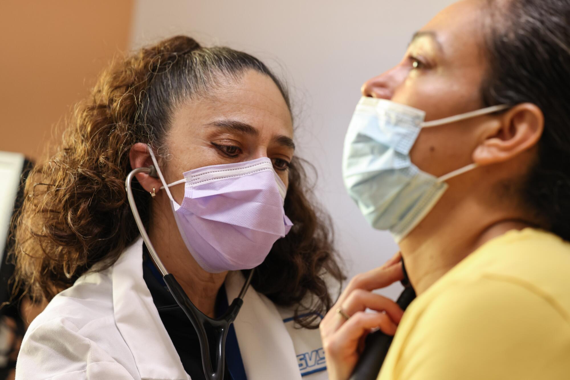 A masked doctor with wavy brown hair holds a stethoscope to the throat of a patient, also masked
