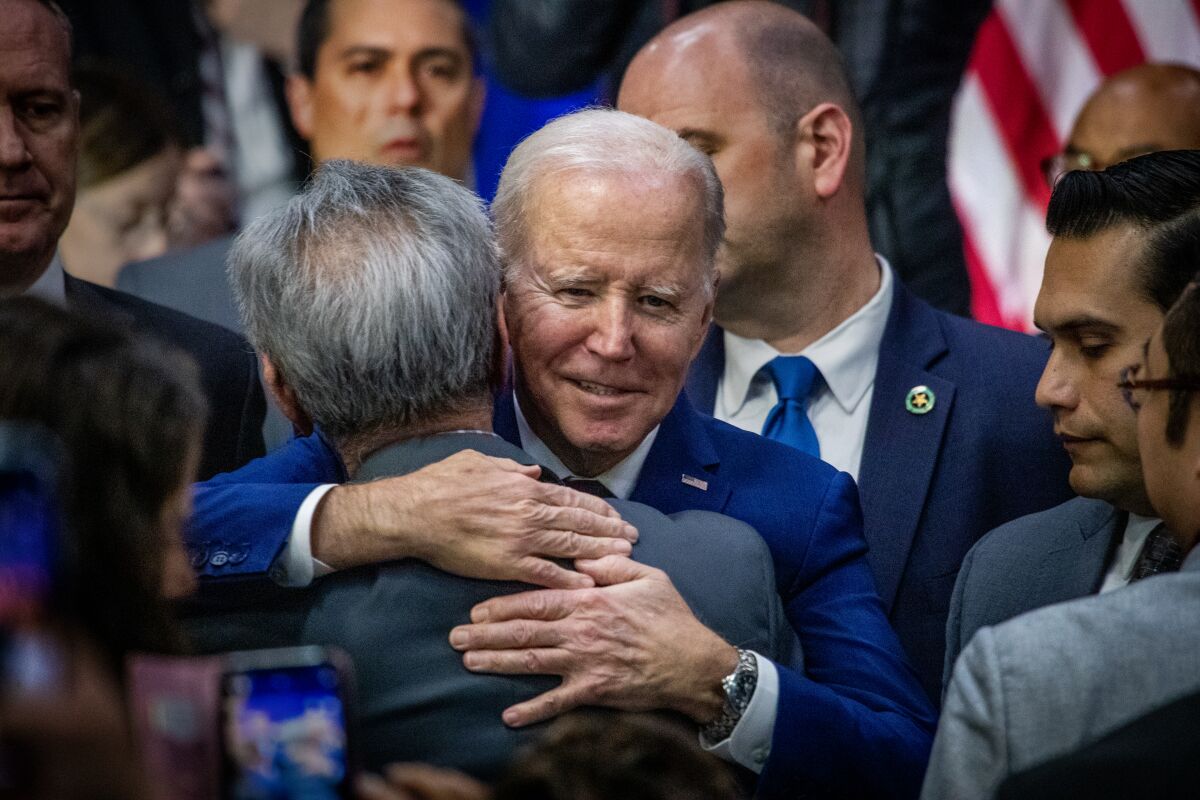 A man with gray hair, in a blue suit, hugs another man as they are surrounded by other people. 