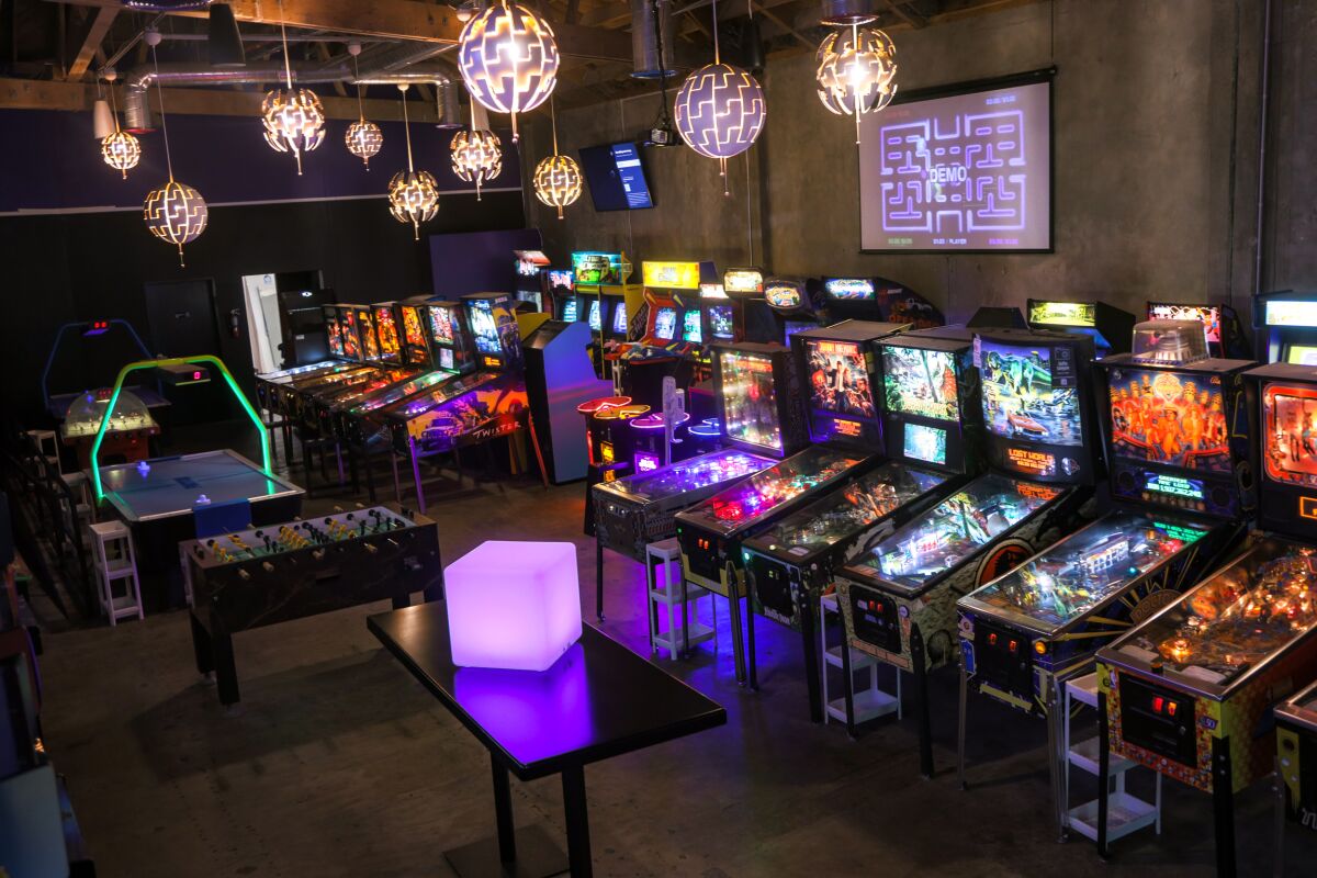 Brightly lighted pinball machines line a wall in a dark room.
