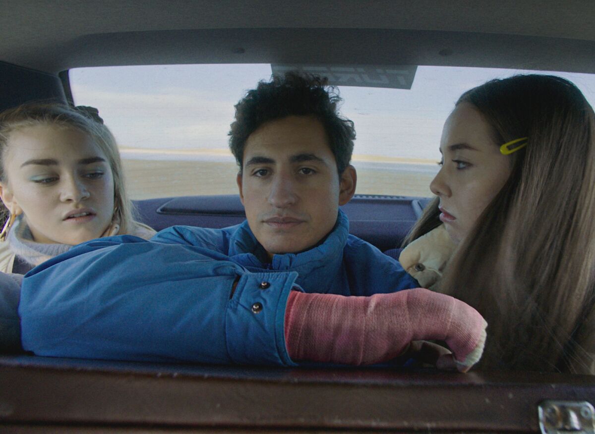 A young man wearing a pink cast sits between two girls in the backseat of a car.