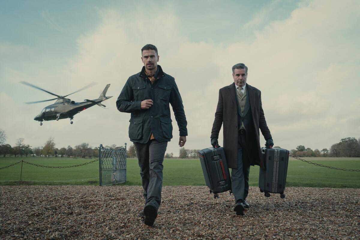 Two men walk on a gravel path and a helicopter takes off behind them.