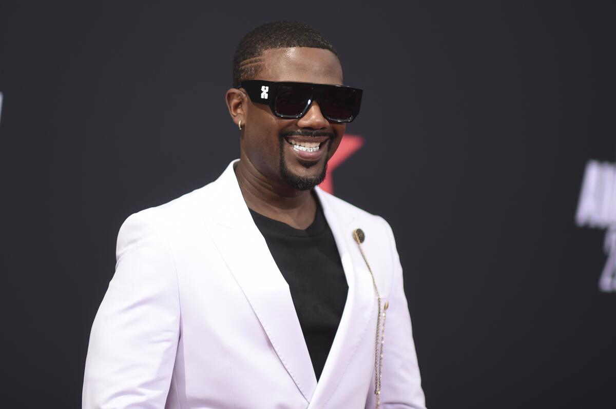A man smiling wearing sunglasses and a white suit.