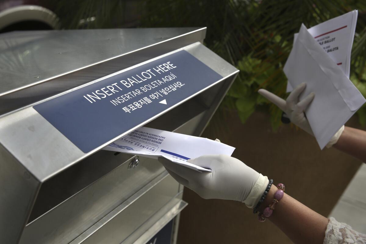 A gloved hand inserts a mail-in ballot envelope into a metal box below a sign that reads "Insert ballot here"