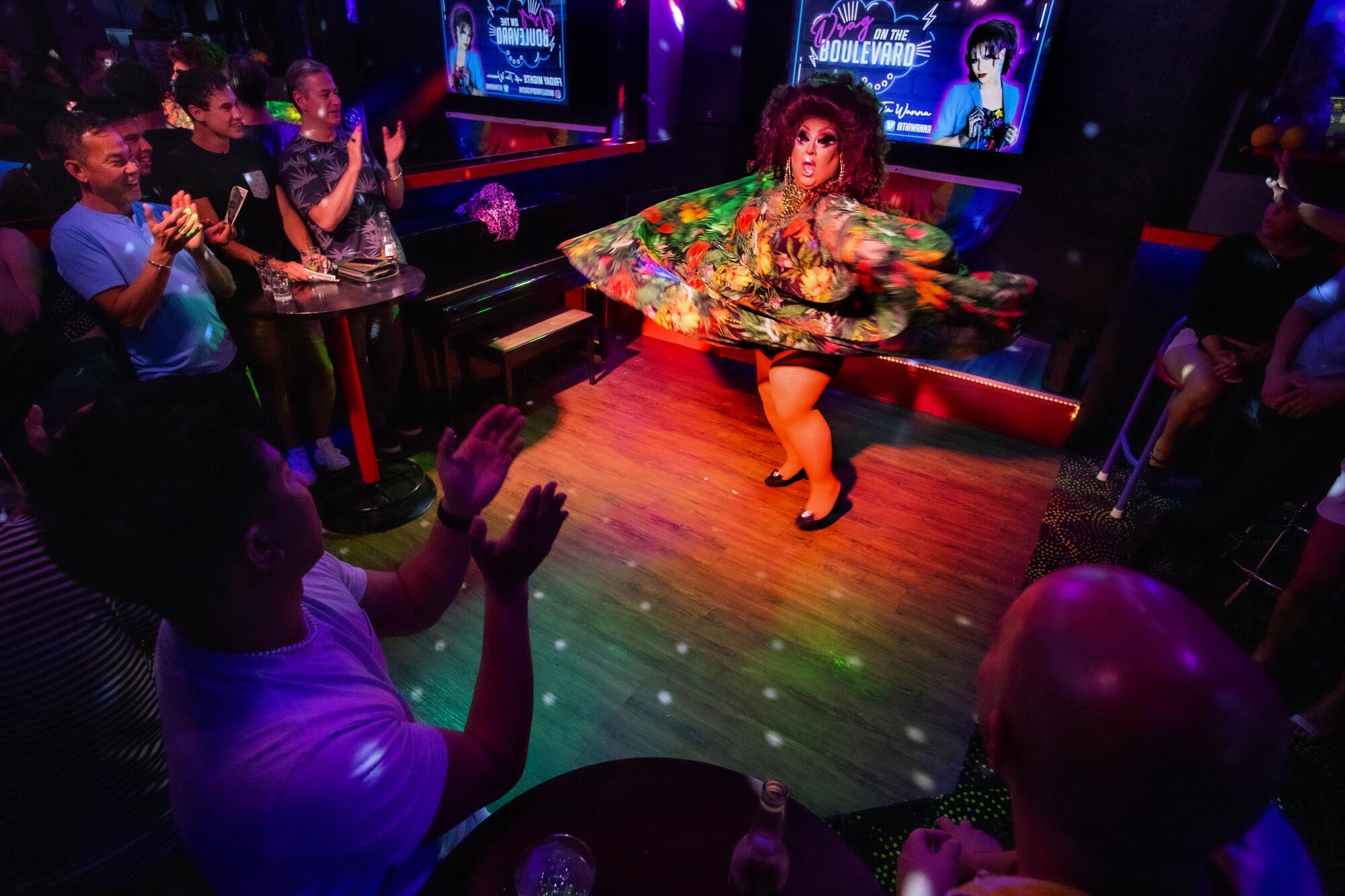 A drag queen performs on a small stage, surrounded by a clapping audience.