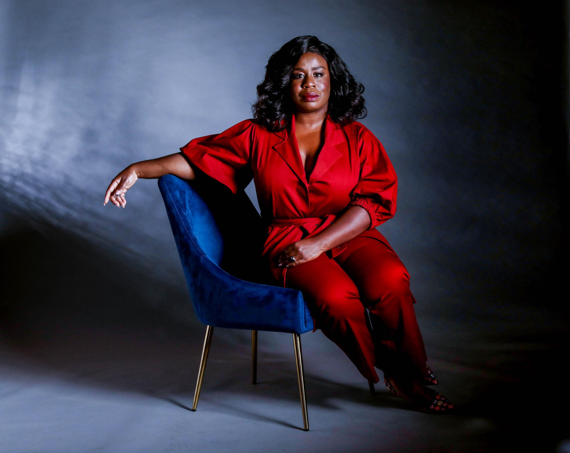 Uzo Aduba, in a red pantsuit, sits on a chair in a studio portrait.