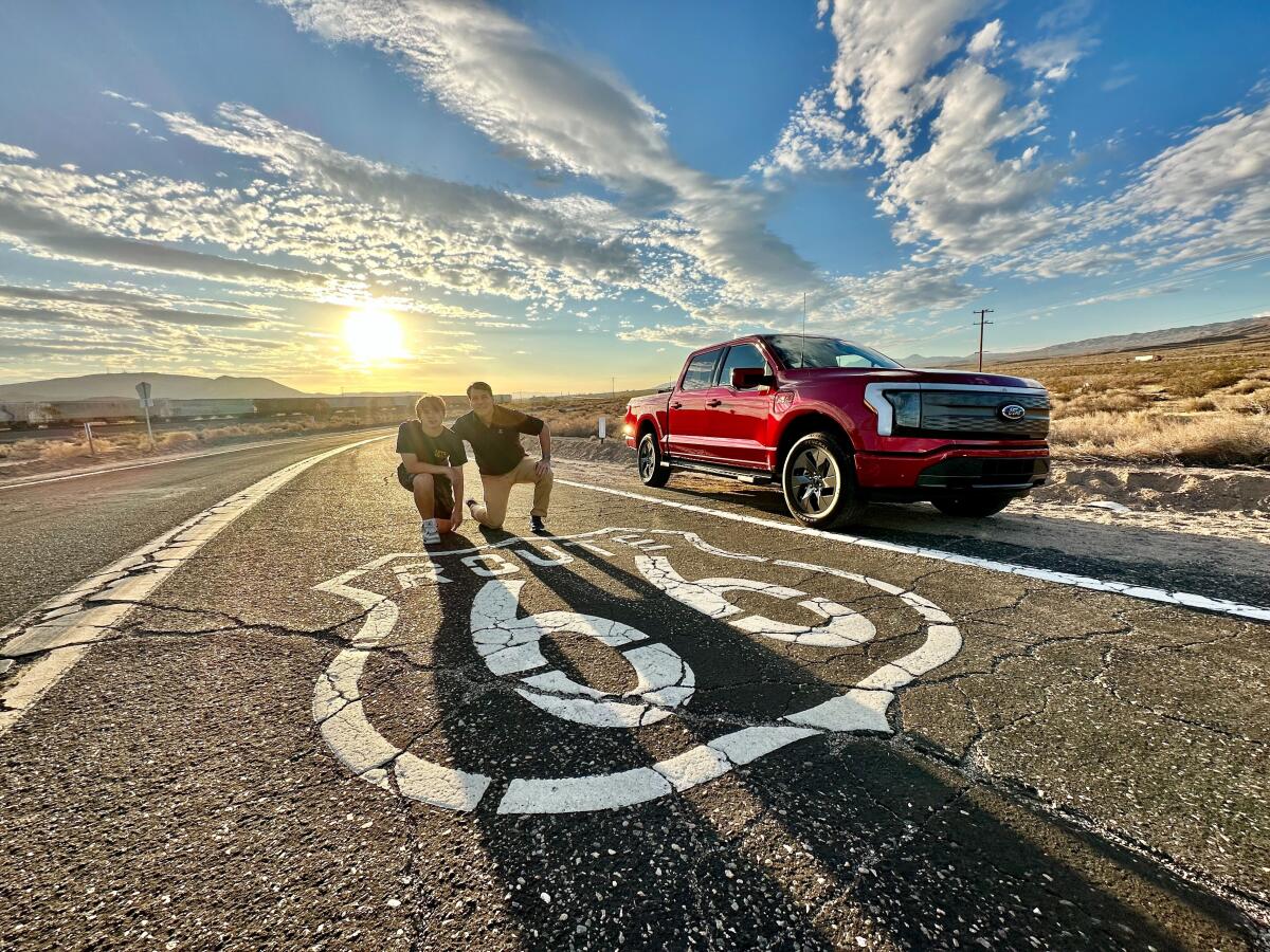 Ford CEO Jim Farley and son kneel on pavement. Red pickup on right. Route 66 sign painted in white on highway.