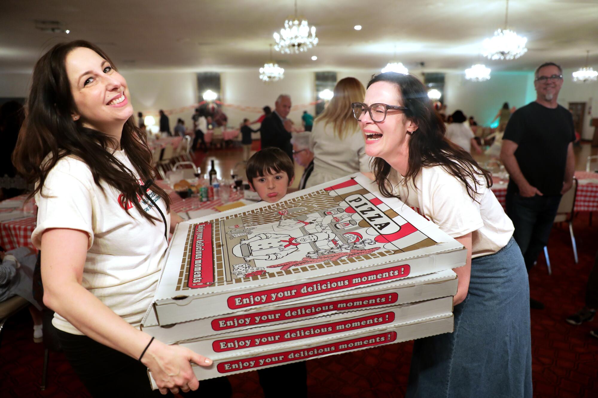 Two women carry large pizza boxes between them, laughing.