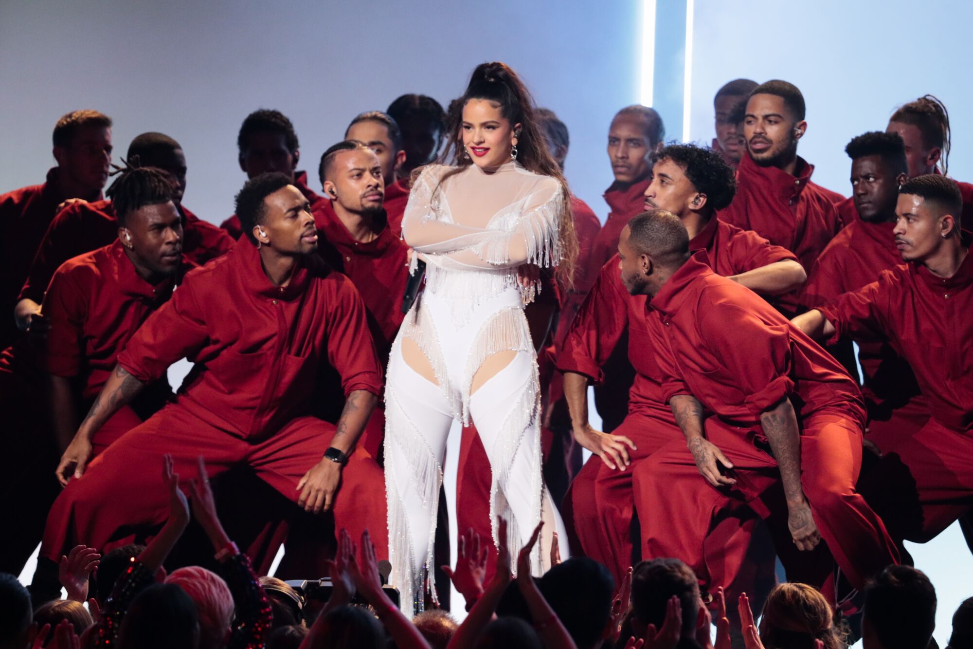 A woman in white performs onstage surrounded by men in red tracksuits.