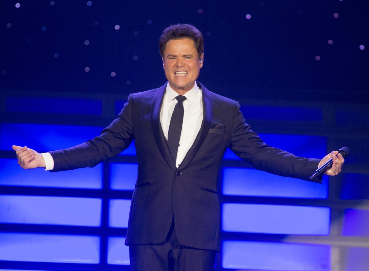 Donny Osmond on stage wearing a suit and tie smiling with his hands out to either side