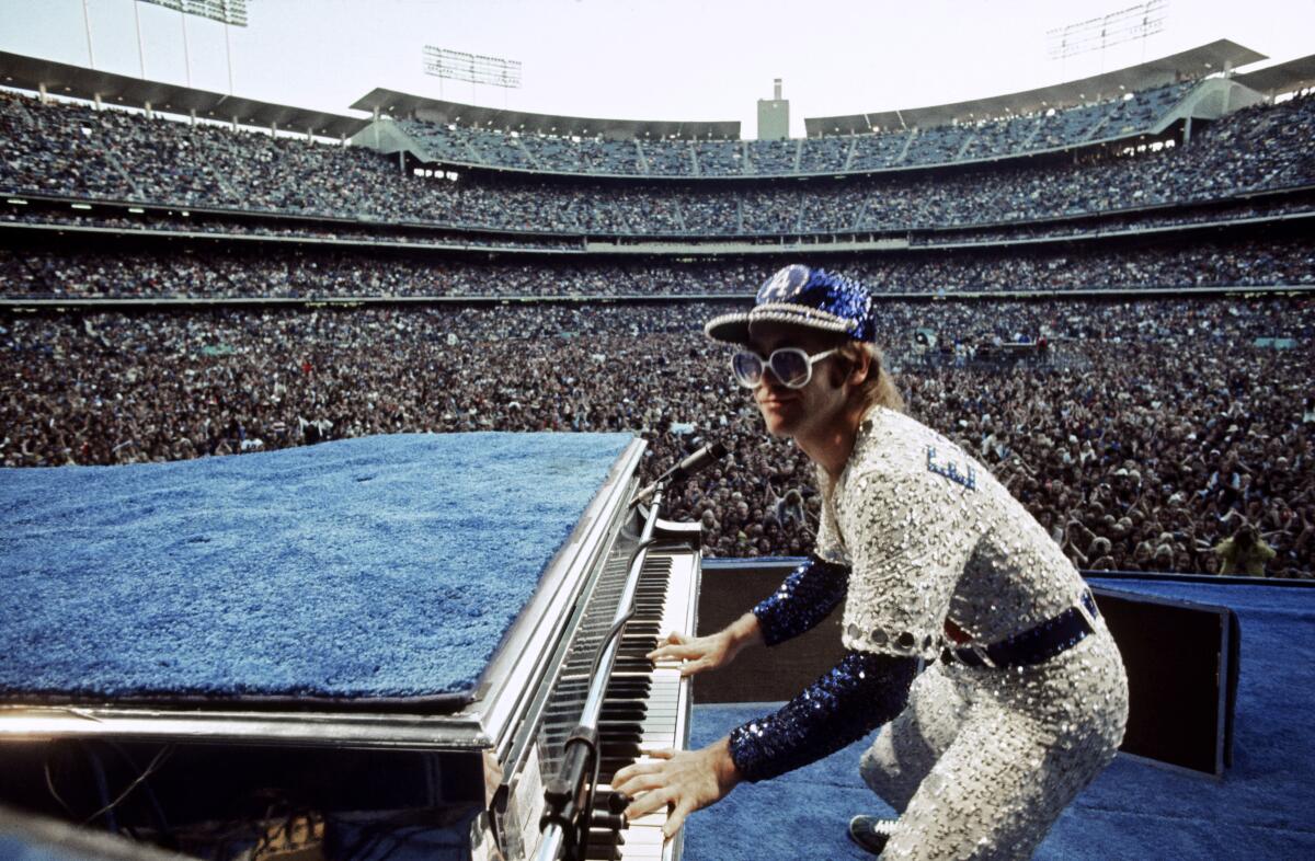 Elton John, dressed in a sequined baseball outfit, performs with the Dodger Stadium crowd behind him.