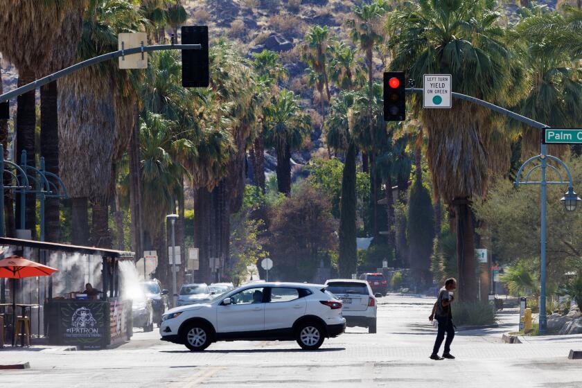 PALM SPRINGS, CA - JULY 8, 2024: A pedestrian clutches his drink as he crosses the street while a customer sits at a local street bar under cooling misters as the temperature reaches 120 degrees on July 8, 2024 in downtown Palm Springs, California.(Gina Ferazzi / Los Angeles Times)