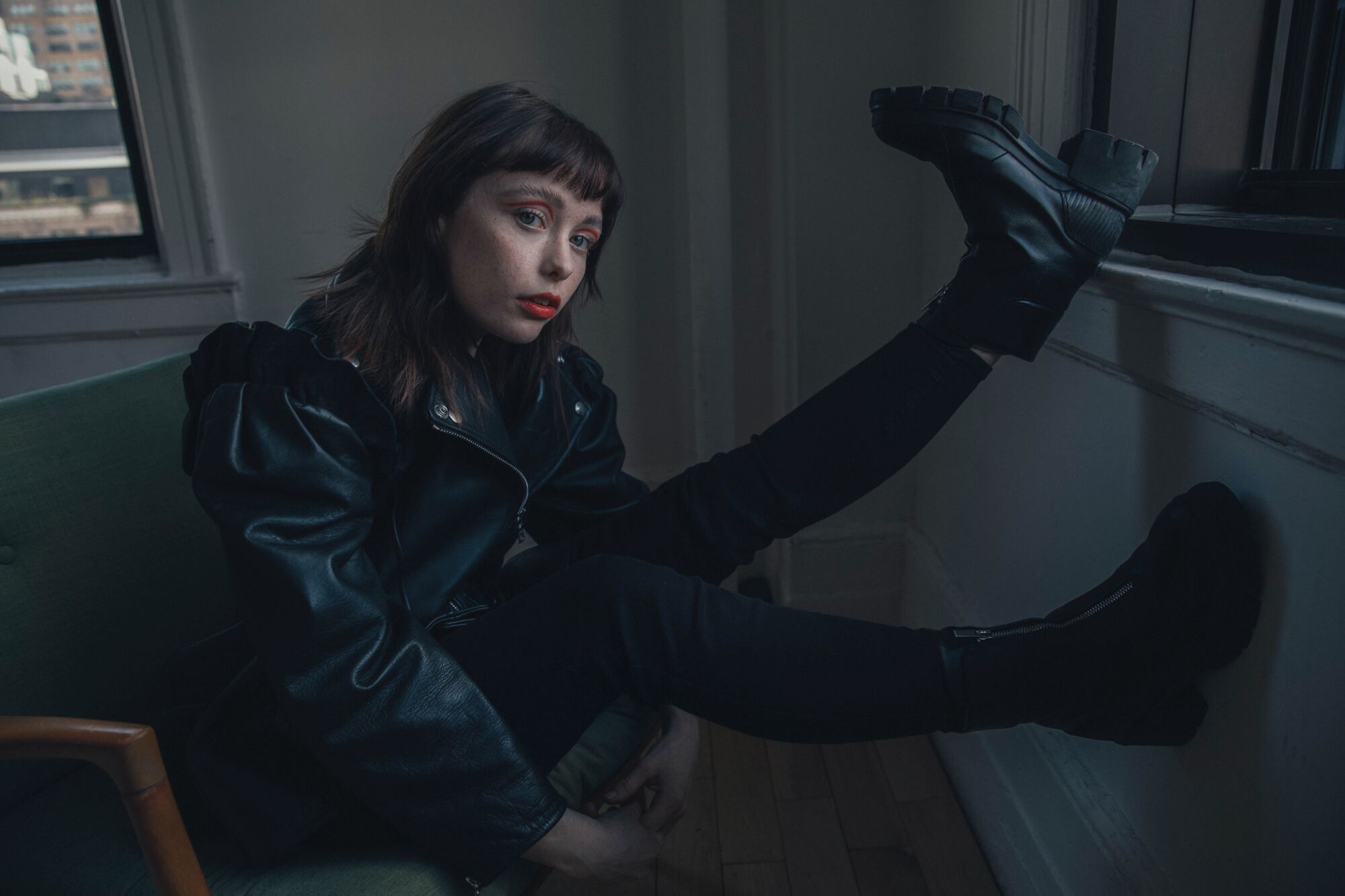 A woman in a leather jacket sits on a chair with her combat boots propped up on a wall.