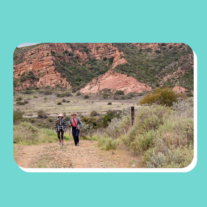 Two hikers along a dirt trial with a reddish mountain range as a backdrop.