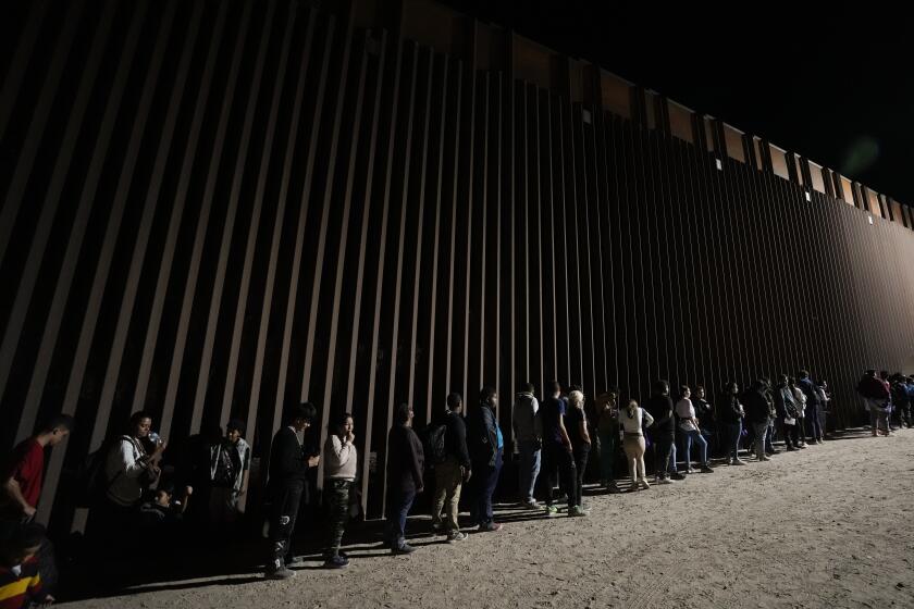 ARCHIVO - Un grupo de personas forman una fila junto a un muro fronterizo mientras esperan para solicitar asilo tras cruzar la frontera desde México, el 11 de julio de 2023, cerca de Yuma, Arizona. (AP Foto/Gregory Bull. Archivo)