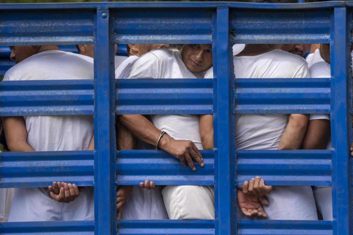 Men wearing white shirts and pants, some handcuffed, look out from behind blue latticed bars 