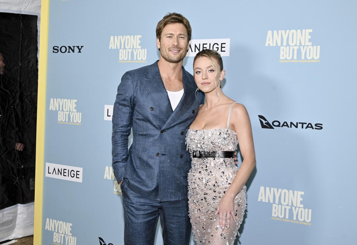 Actor Glen Powell in a blue-gray suit posing with one arm around Sydney Sweeney, clad in a belted, sparkly, strappy dress