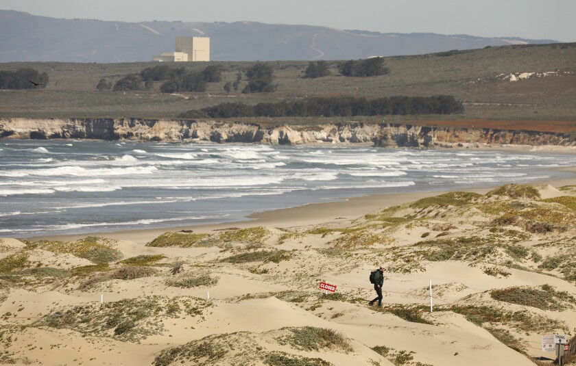 A man walks along a beach as waves roll in. One building is seen in the distance.