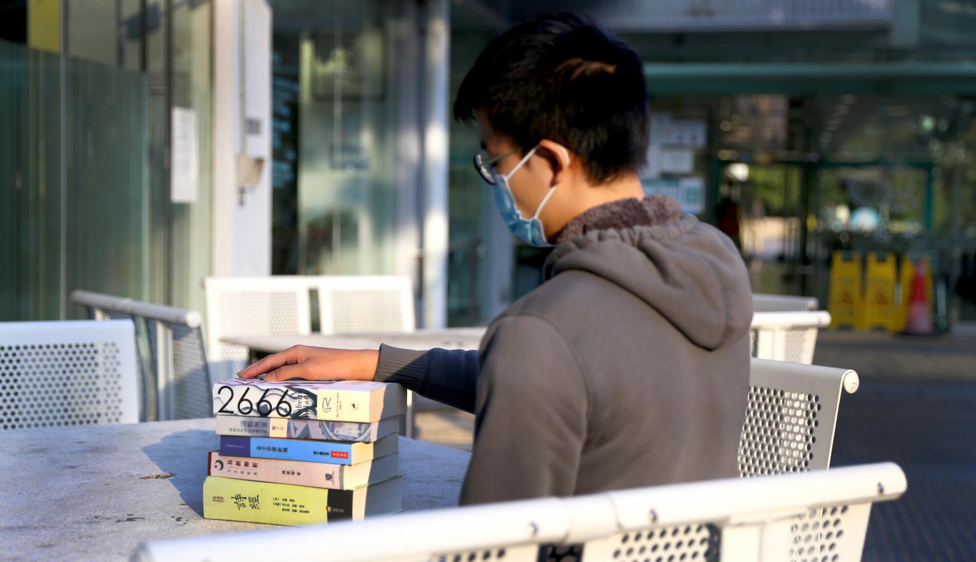 A student sits at a table with a stack of books in front of him.