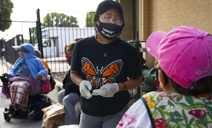 A young woman in a mask that says Community wellness is key works at an outdoor food relief station