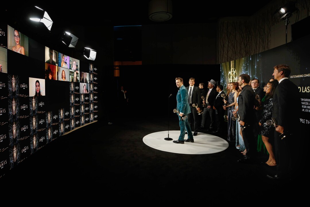 A man in a blue-green suit holds a statuette. Behind him is a line of people holding similar statuettes