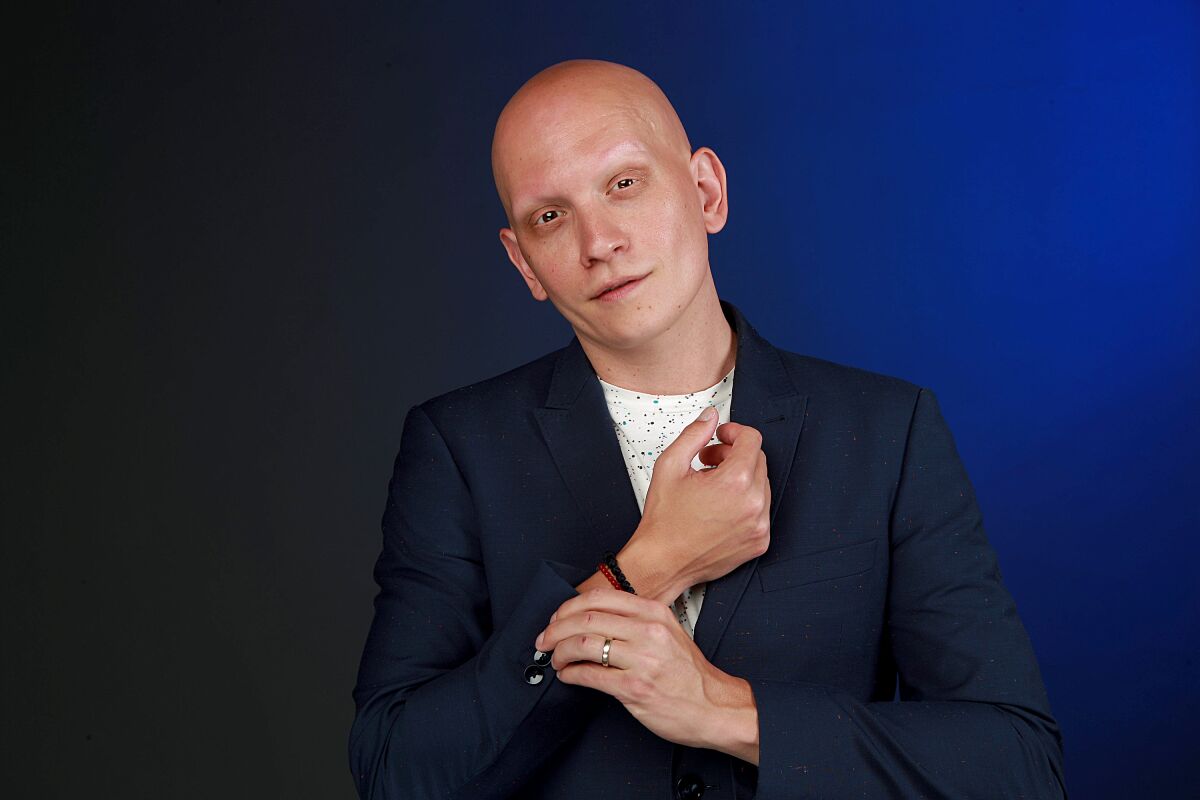 Anthony Carrigan in a t-shirt and blazer, holding his wrist