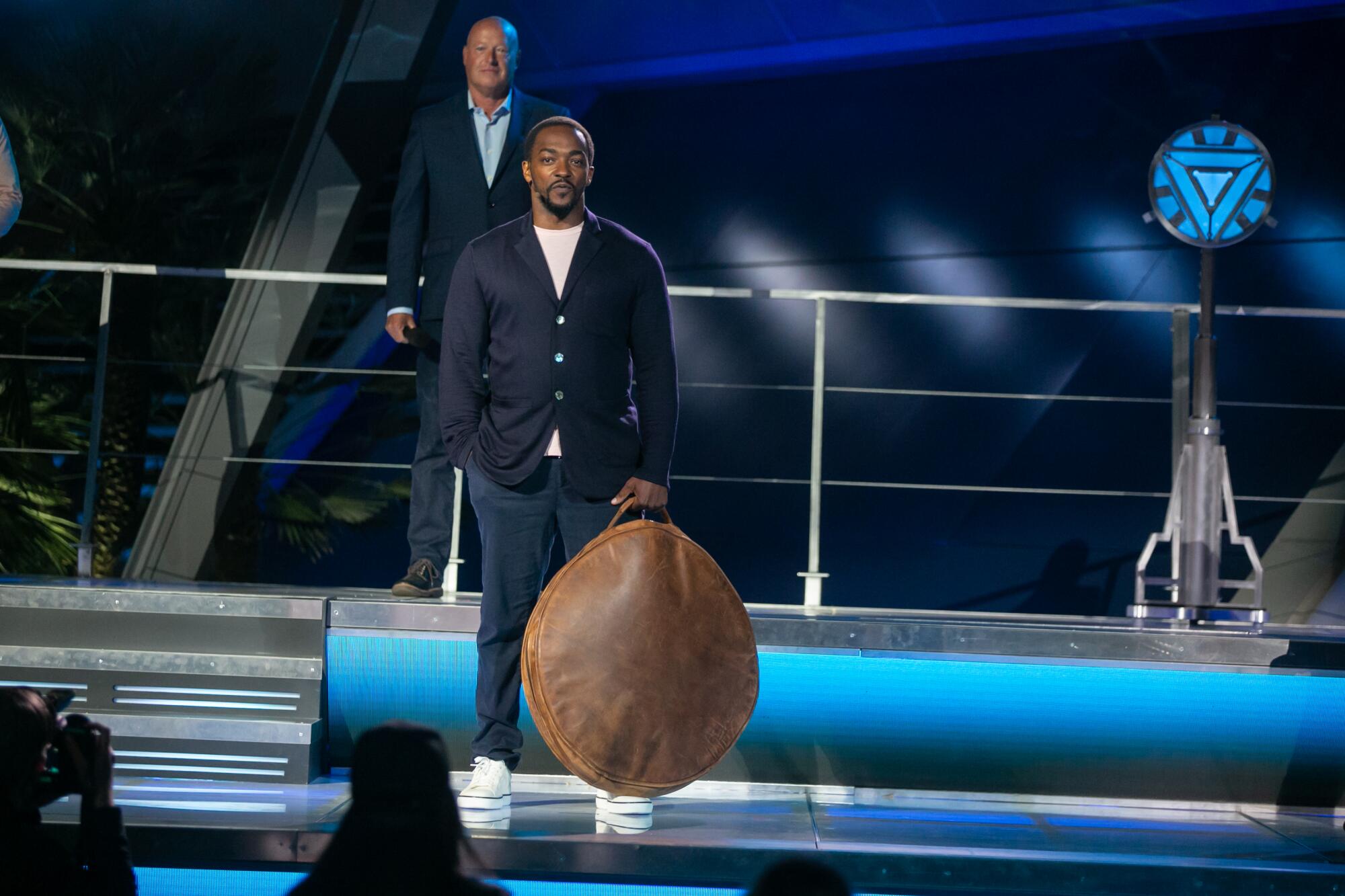 Anthony Mackie, in a dark jacket and white tennis shoes, holds a large round bag.