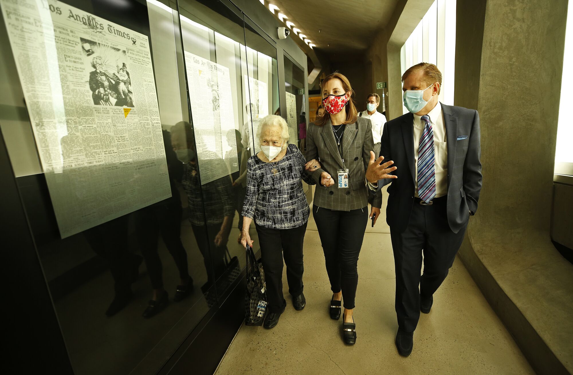 Three people walk down a hallway whose walls are lined with framed newspaper pages