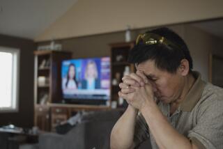 A man with his head bowed and his hands clasped with a television on in the background in the documentary "Bad Axe."