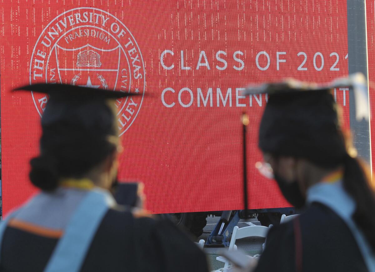 Two students in caps and gowns facing a red backdrop at a University of Texas graduation ceremony