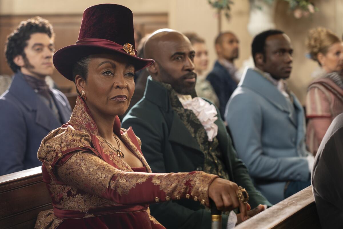 Actors Adjoa Andoh in a hat and dress and Daniel Francis in a period jacket sitting in a pew in a scene from "Bridgerton"