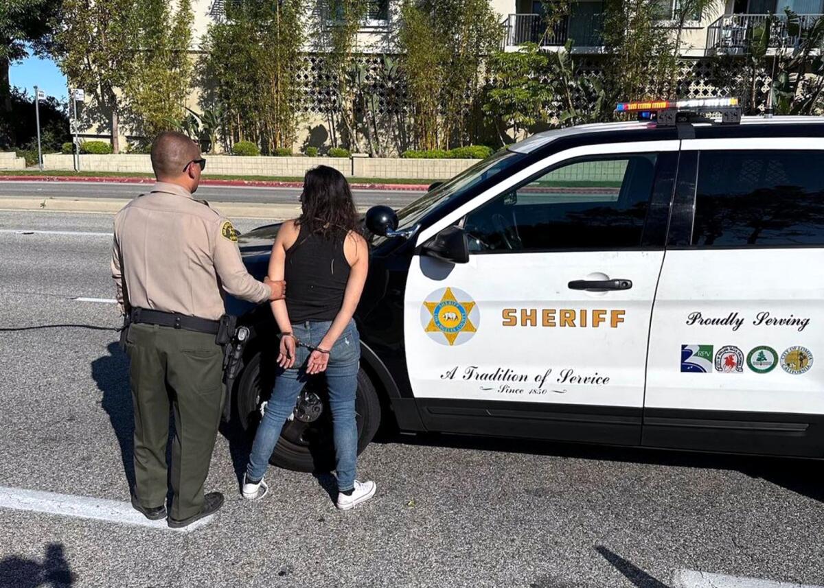 A sheriff's deputy holds on to an arm of a person handcuffed while standing next to a patrol car.