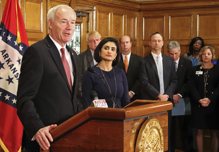 Gov. Asa Hutchinson, left, speaks at a lectern while several people in a wood-paneled room look on.